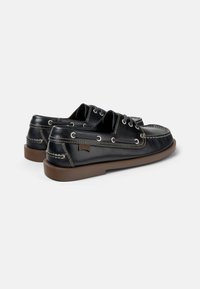 Zapatos de barco de cuero negro con suela de goma marrón, detalles de costura, ojales de metal y logo en el lateral. Diseño clásico y perfil bajo.