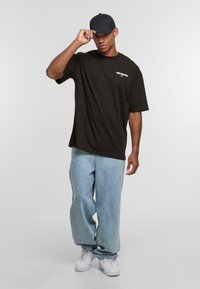 Black oversized t-shirt with logo on the left chest, paired with light blue baggy jeans and white sneakers. Black cap worn tilted.