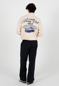 Pull en maille beige avec texte graphique bleu et illustration de voiture au dos. Associé à un pantalon noir ample et des chaussures de couleur claire.