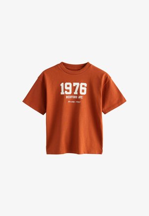 Oranžové bavlněné tričko s krátkým rukávem. Na přední straně se nachází bíle tištěný text "1976 BEDFORD AVE" a "Brooklyn, NYC".