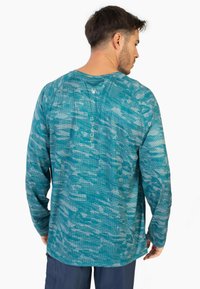 Camicia a maniche lunghe in teal con un motivo camouflage, realizzata in tessuto textured. Presenta un logo sulla schiena e un collo a giro.