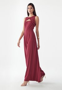 Femme portant une combinaison sans manches bordeaux avec jambes larges plissées, décolleté découpé, bracelet doré et sandales à talons hauts dorées, debout sur un fond blanc.