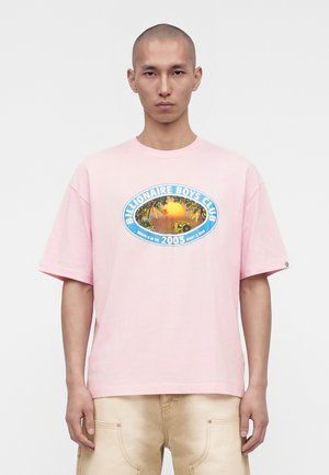 Homme aux cheveux courts portant un t-shirt rose Billionaire Boys Club avec un motif coucher de soleil et palmier sur le devant, pantalon beige visible.