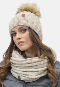 Caciula bej tricotată cu un pompon din blană și o eșarfă tricotată infinity asortată. Ambele articole au un model texturat și o etichetă mică din piele.
