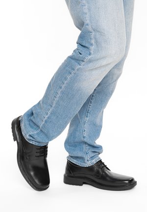 Person wearing light blue jeans and black polished leather dress shoes, standing with one leg crossed over the other.