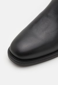 Botín de cuero negro hasta el tobillo con punta redondeada, textura suave y un brillo sutil. Presenta costuras simples y una suela plana de goma.