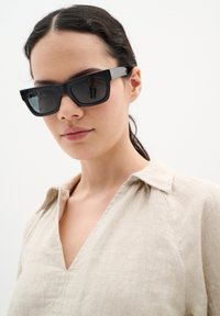 Čierne oversized slnečné okuliare s geometrickým tvarom, majúce výrazný rám a tmavé sklá. Nosené s béžovou ľanovou košeľou s golierom.