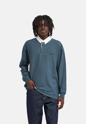 Jonge man met halflange dreadlocks staat met één hand in zijn zak, gekleed in een blauw gestreepte longsleeve polo en donkere jeans.