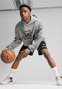 Haut à capuche gris avec le logo "PUMA" noir, shorts de sport noirs et baskets blanches avec des détails rouges. Tenant un ballon de basket, dans une pose dynamique.