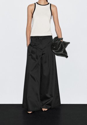 Femme portant un haut blanc sans manches bordé de noir, un pantalon noir à jambes larges, tenant une pochette noire texturée.