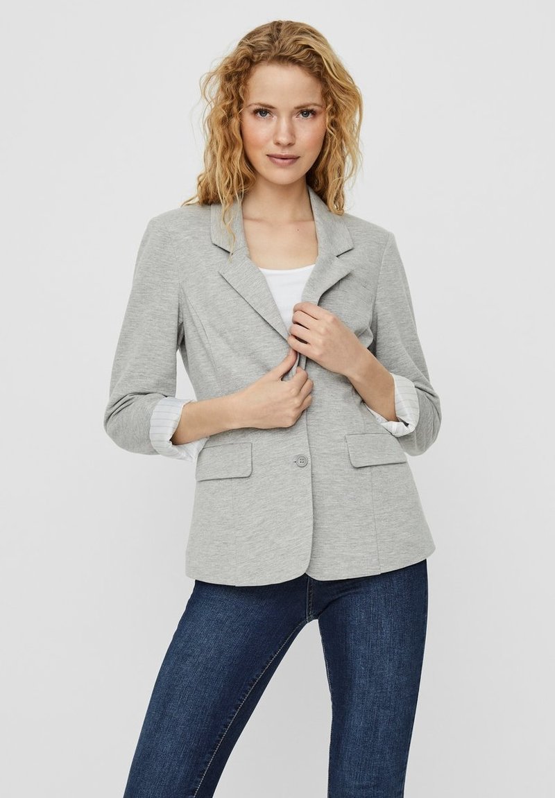 Blazer gris clair en tissu doux, avec un col cranté, deux poches latérales et une fermeture à un bouton. Porté sur un haut blanc.