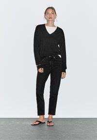 Top nero a maniche lunghe con scollatura a V, sovrapposto a una camicia bianca, abbinato a jeans neri a gamba dritta e ciabatte nere, su uno sfondo uniforme.