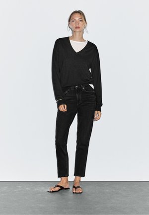 Czarna koszulka z długim rękawem w serek nałożona na białą koszulę, w zestawie z czarnymi prostymi dżinsami i czarnymi klapkami, na jednolitym tle.