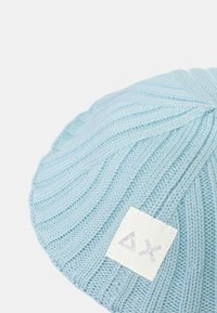 Cappello a maglia azzurro chiaro con texture a coste e etichetta del logo triangolare bianca con accenti argento, caratterizzato da una forma arrotondata.