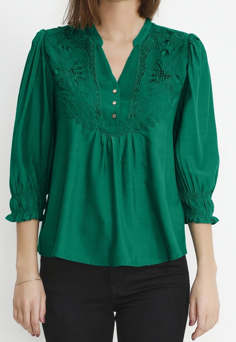 Blouse verte avec encolure en V, broderie décorative sur le corsage, boutons décoratifs et manches bouffantes élastiquées, confectionnée dans un tissu lisse.