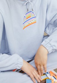 Felpa azzurro chiaro con un grande logo grafico colorato che presenta archi arcobaleno e testo. Polsini a coste e dettaglio con cordino. Vestibilità casual.