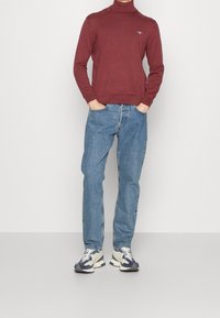 Burgunderfarbener Rollkragenpullover, Baumwollmischung, körpernahe Passform. Blaue Jeans, gerade geschnitten, helle Waschung. Sneaker in Creme- und Marineblau.