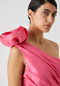 Pink silk dress featuring a draped design and an oversized floral accent on the shoulder. Smooth texture with a subtle sheen.