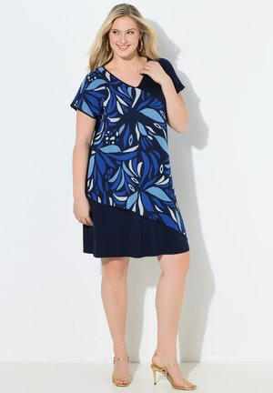 Kobieta ubrana w granatową sukienkę z krótkim rękawem z asymetrycznym niebieskim wzorem liści, złote kolczyki koła i złote sandały na wysokim obcasie.