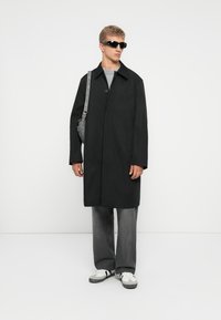 Abrigo largo negro con cierre de botones y un cuello estructurado, llevado sobre una camisa gris. Los accesorios incluyen un bolso gris y gafas de sol.