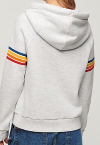 Jasnoszara bluza z kapturem ze sznurkiem i wielokolorowymi paskami (czerwonym, żółtym, niebieskim) na rękawach; miękka tekstura i ściągacz na dole.