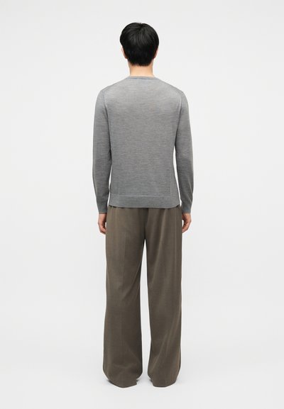 Muž stojící zády, oblečený v dlouhém šedém svetru a volných hnědých kalhotách, na jednobarevném bílém pozadí.