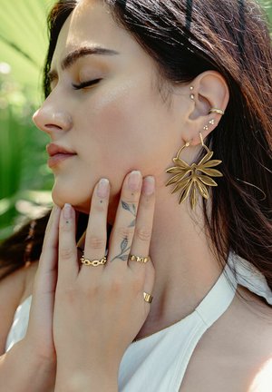 Gold leaf-design earrings, multiple gold rings, and a simple white top. Smooth textures with a mix of intricate details. Natural light background.
