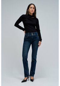 Top negro de manga larga con un detalle drapeado, combinado con unos pantalones vaqueros ajustados azules oscuros. La modelo lleva unos zapatos de tacón negros. Fondo sencillo.