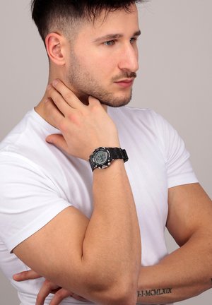 Orologio digitale con quadrante rotondo nero, marcatori delle ore in grassetto e cinturino in gomma testurizzata. Indossato su un polso, in evidenza con una maglietta bianca casual.