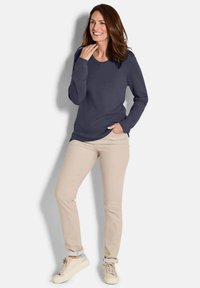 Frau mit langen braunen Haaren, die lächelt und ein navyblaues Langarmshirt, beige Hosen und hellbeige Sneaker trägt, steht vor einem weißen Hintergrund.