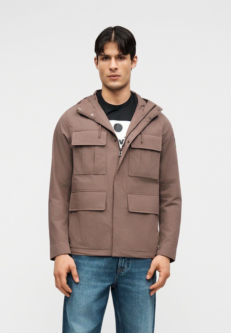 Jeune homme portant une veste à capuche marron avec quatre poches à rabat à l'avant et un jean bleu, debout devant un fond clair uni.