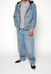 Ljusblå denimjacka med huva, grå t-shirt, lösa ljusblå jeans och svarta sneakers med vit sula och rutiga detaljer.