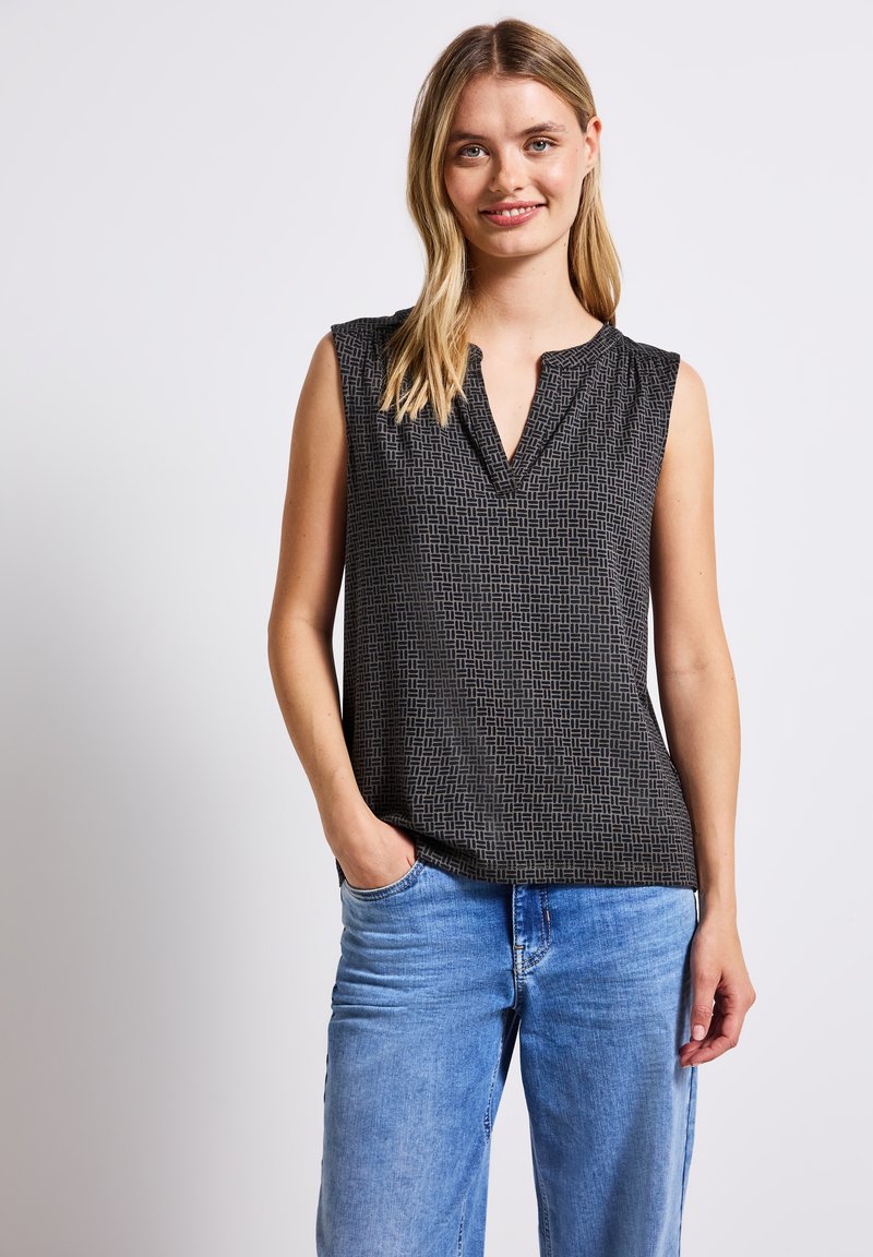 Ärmlös svart topp med geometriskt mönster, v-ringning och subtila veckdetaljer, tillsammans med ljusblå jeans. Tyget är en bomullsblandning.