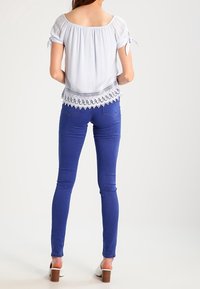 Blusa azzurra chiara con spalle scoperte, con lacci regolabili e bordo in pizzo, abbinata a jeans skinny blu e sandali con tacco bianchi.