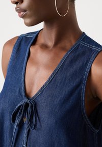 Gros plan d'une personne portant un haut sans manches en denim bleu foncé avec un nœud à l'avant et une grande boucle d'oreille.