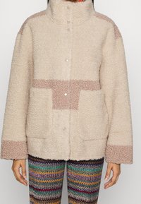 Flauschige beige Jacke mit hohem Kragen, ausgestattet mit hellbraunen Akzenten, großen Taschen und Druckknöpfen; getragen über mehrfarbigen Zickzack-Musterhosen.