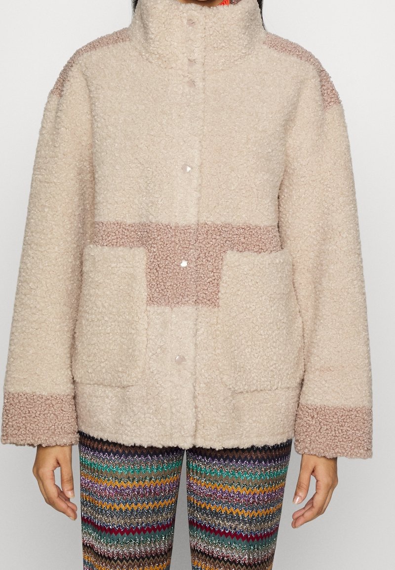 Flauschige beige Jacke mit hohem Kragen, ausgestattet mit hellbraunen Akzenten, großen Taschen und Druckknöpfen; getragen über mehrfarbigen Zickzack-Musterhosen.