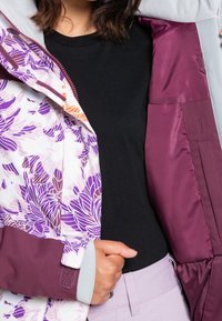 En person som bär en lila blommig jacka, öppen för att avslöja en enfärgad svart skjorta och ljuslila byxor, håller jackans kant med ena handen.