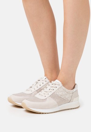 Woman wearing beige and white Michael Kors sneakers with lace-up design and logo pattern on the heel and side panels.