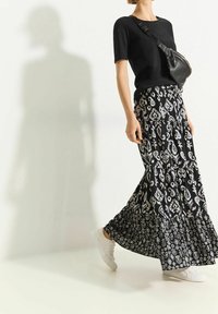 T-shirt noire associée à une jupe longue à volants noire et blanche à motifs, portée avec des baskets blanches. La tenue est complétée par un sac bandoulière.