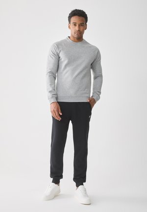 Man standing, wearing a light gray long-sleeve sweatshirt, black jogger pants, and white sneakers against a plain background.