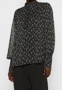 Blouse noire à manches longues, col haut et devant boutonné. Présente un motif répétitif de formes abstraites en blanc et teal. Tissu doux.