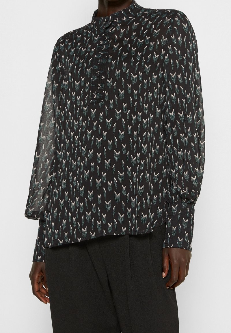 Blouse noire à manches longues, col haut et devant boutonné. Présente un motif répétitif de formes abstraites en blanc et teal. Tissu doux.