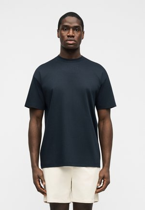 Mannlig modell står stille iført en ensfarget mørk marineblå kortermet skjorte og lyse beige shorts mot en hvit bakgrunn.