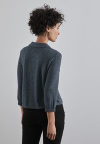 Mörkblå stickad topp med krage, trekvartslånga ärmar och elastiska ärmslut, bärs över svarta byxor. Texturerad tygdetalj synlig.