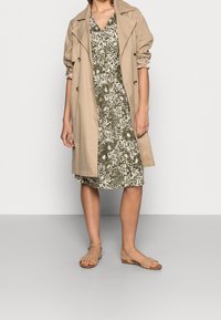 Beige trenčkot přes zelené květinové šaty, ukazující výstřih do V a krátké rukávy. Oblečení doplňují béžové ploché sandály.