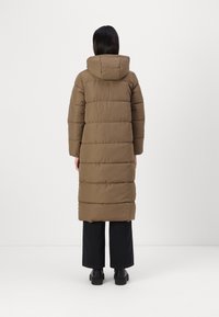Brun pufferjakke med hette, med et quiltet design og lang lengde, kombinert med svarte vide bukser og kraftige svarte støvler.