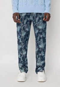 Un homme portant un pantalon en camouflage bleu et gris, un pull en tricot léger bleu clair et des baskets blanches se tenant contre un fond blanc.