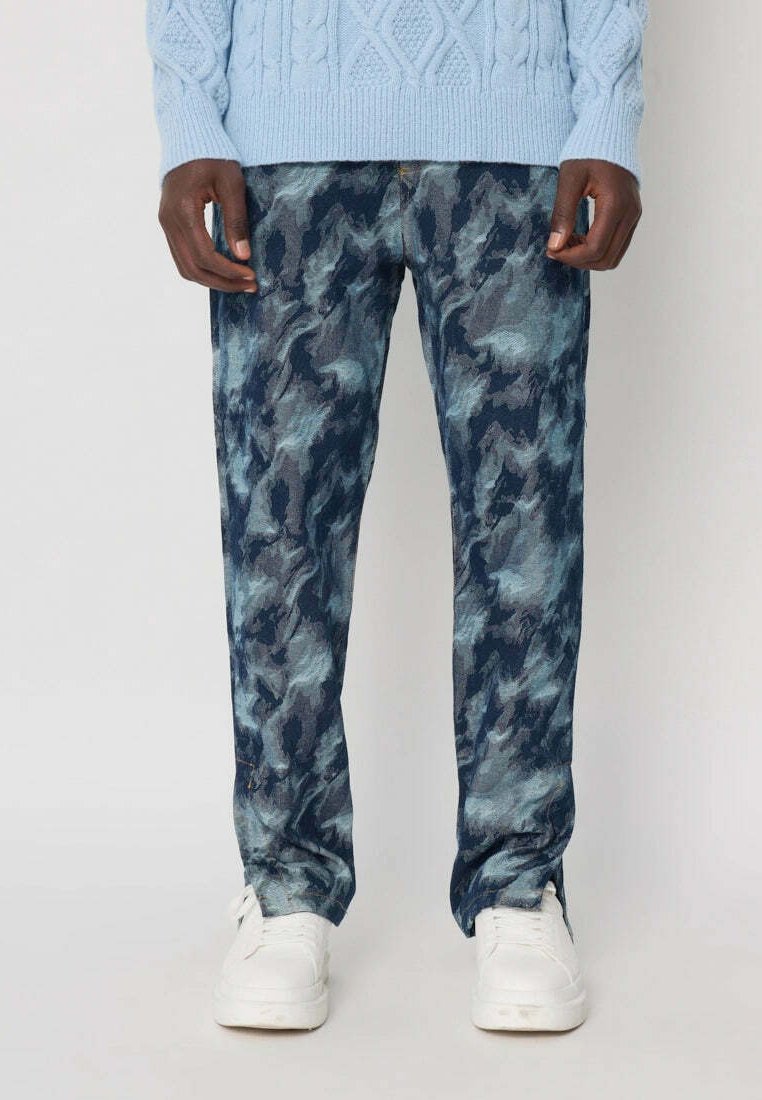 Un homme portant un pantalon en camouflage bleu et gris, un pull en tricot léger bleu clair et des baskets blanches se tenant contre un fond blanc.