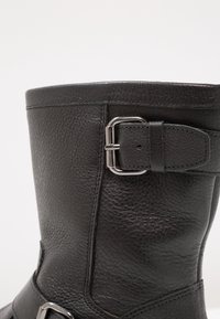 Bota de cuero negro con un acabado texturizado, que presenta un acento de hebilla lateral y detalles de costura para un diseño adicional. Caña alta y punta redondeada.